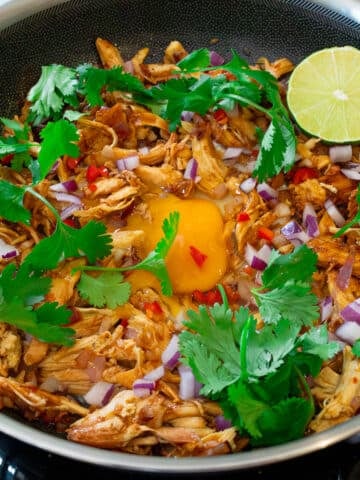 Filipino chicken sisig with shredded rotisserie chicken, a runny egg, red onion, chili, cilantro, and lime in a skillet