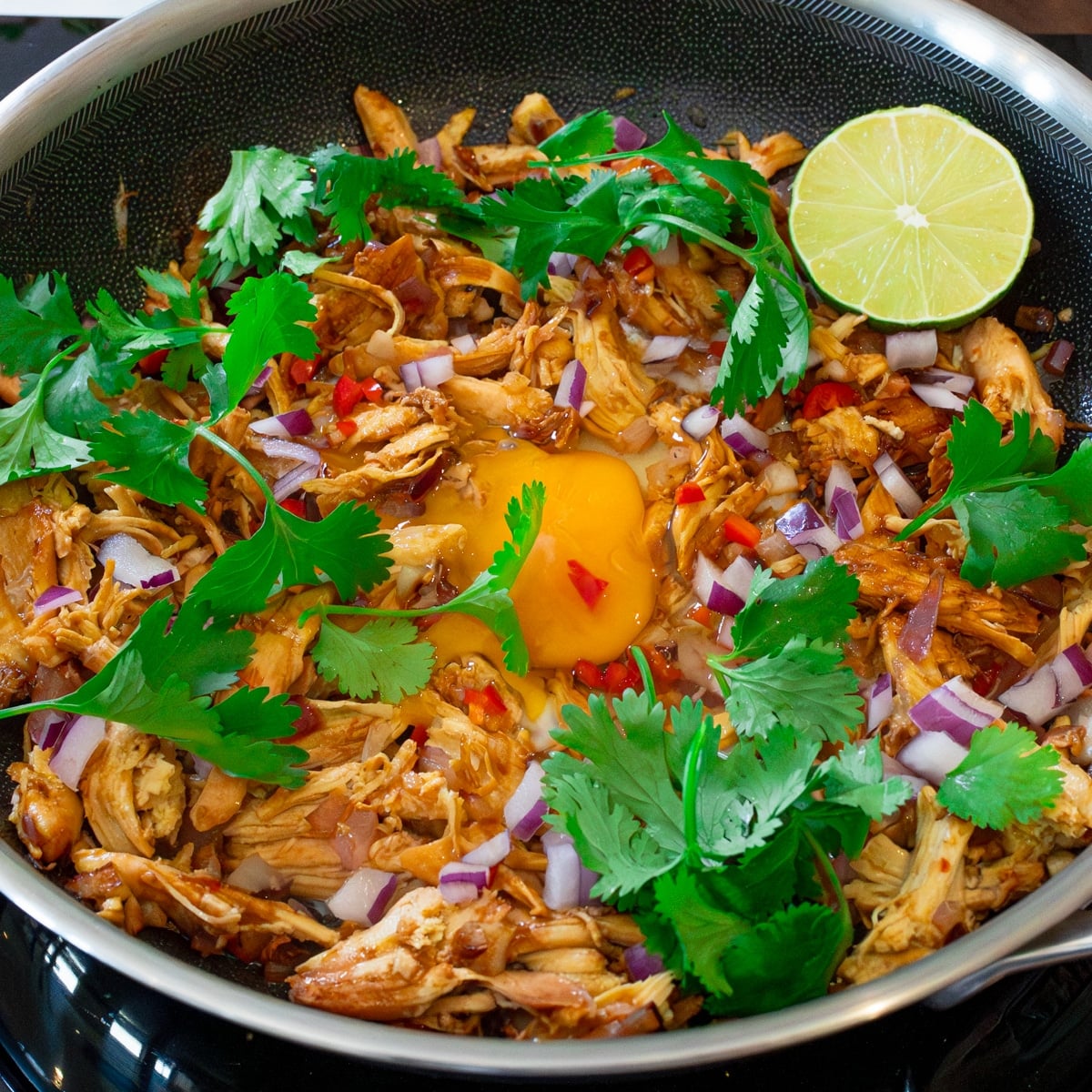 Filipino chicken sisig with shredded rotisserie chicken, a runny egg, red onion, chili, cilantro, and lime in a skillet