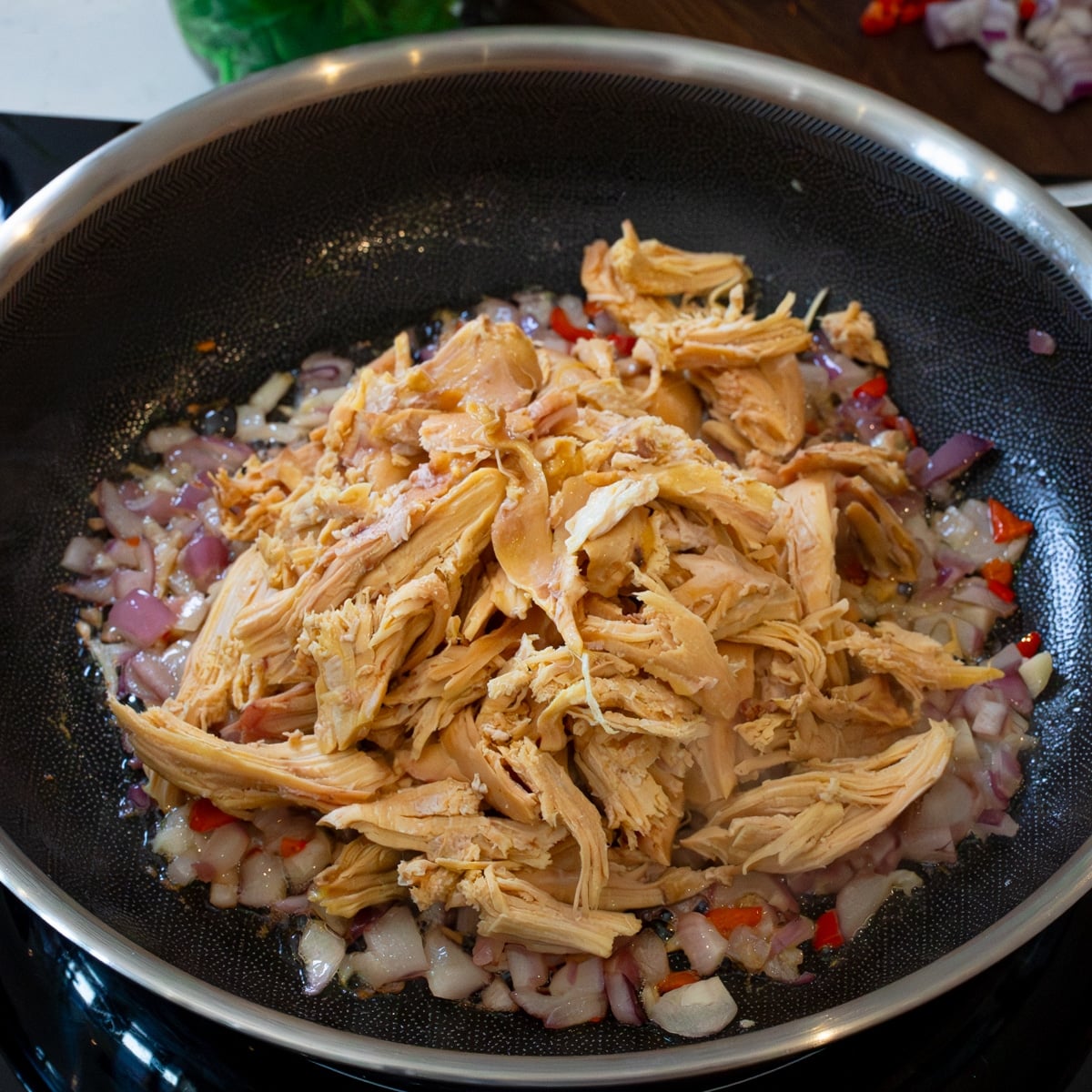 Shredded rotisserie chicken added to onion, garlic, and chili in a pan for chicken sisig