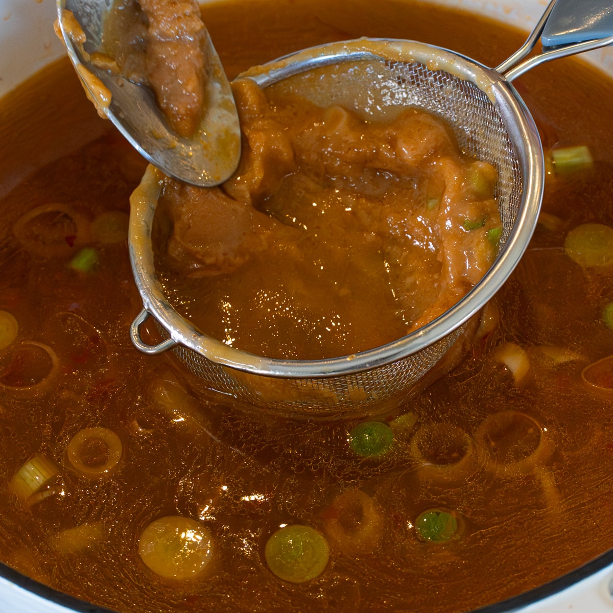 Miso paste being pressed through a fine mesh strainer directly into chicken bone broth for chicken miso ramen