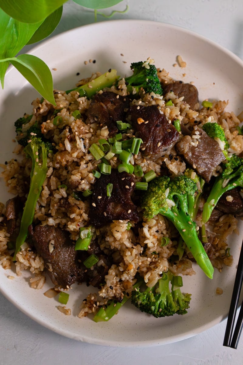 Beef and broccoli fried rice with tender beef strips, broccoli florets, green onions, sesame seeds, and chopsticks on a white plate.