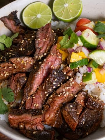 Asian marinated skirt steak bowl with honey sriracha glaze, steamed rice, fresh mango cucumber salsa, lime wedges, sesame seeds, and cilantro
