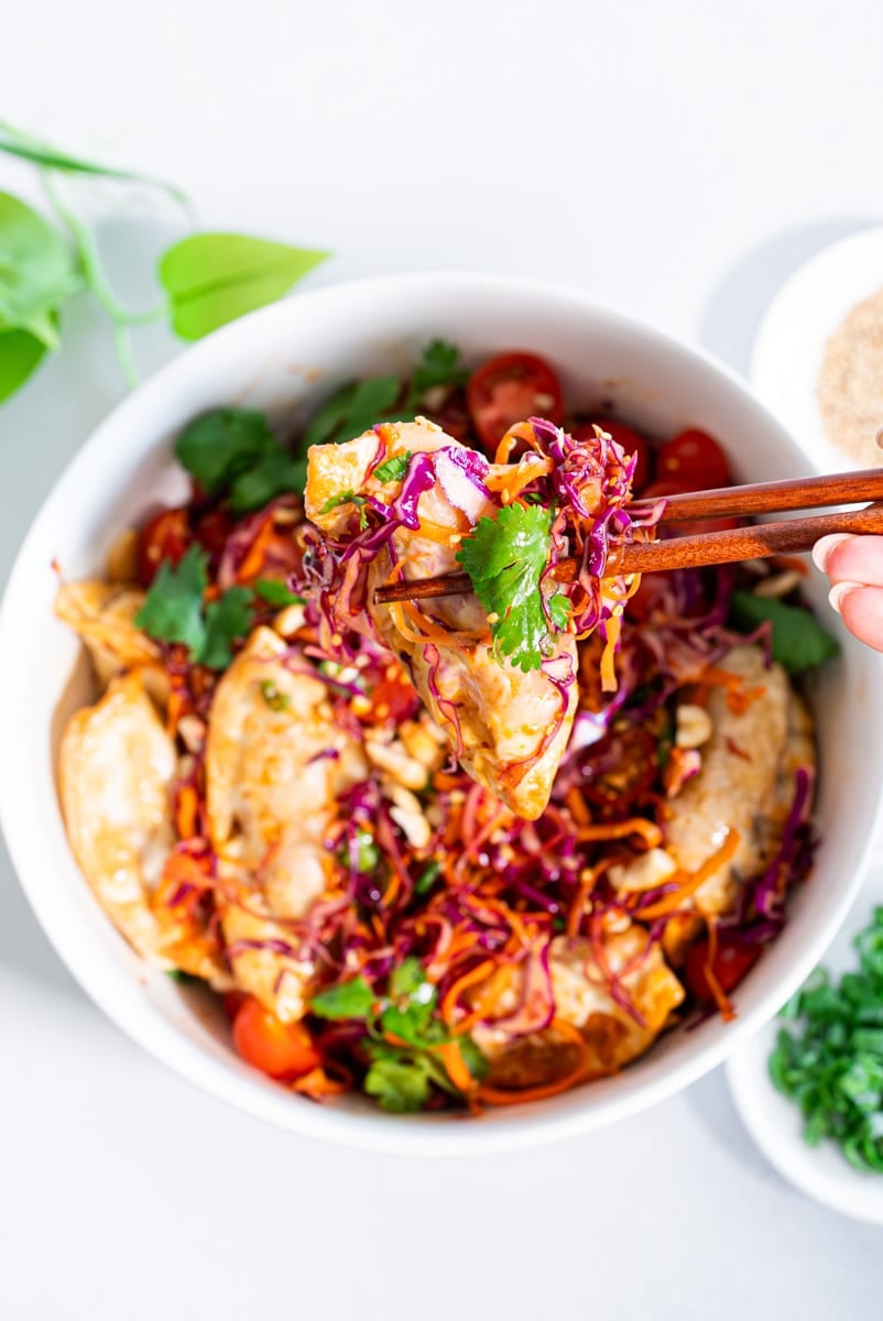 Overhead view of dumpling salad in a white bowl with crispy dumplings, shredded purple cabbage, carrots, cherry tomatoes, cilantro, and cashews, with chopsticks lifting a bite