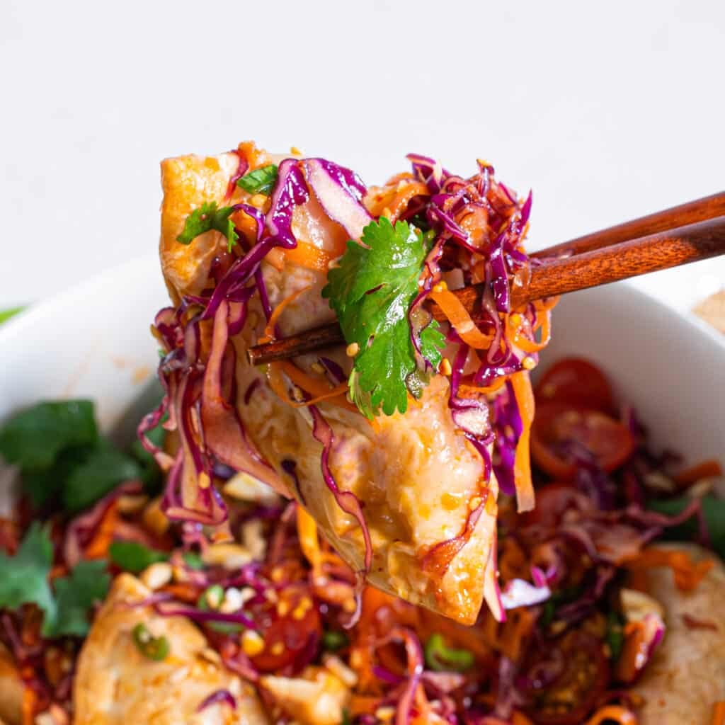 Close-up of a crispy dumpling lifted with chopsticks, topped with shredded purple cabbage, cilantro, and honey soy dressing over a bowl of dumpling salad