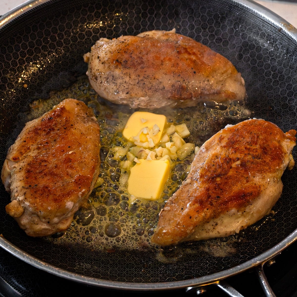 Chicken breasts pan frying in oil with butter and garlic in a HexClad skillet for maple glazed chicken