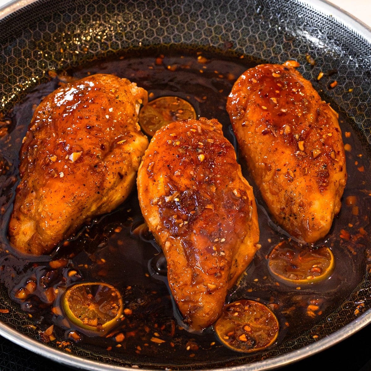 Maple glazed chicken breasts finishing in the sticky soy chili crunch glaze with lime slices in a skillet